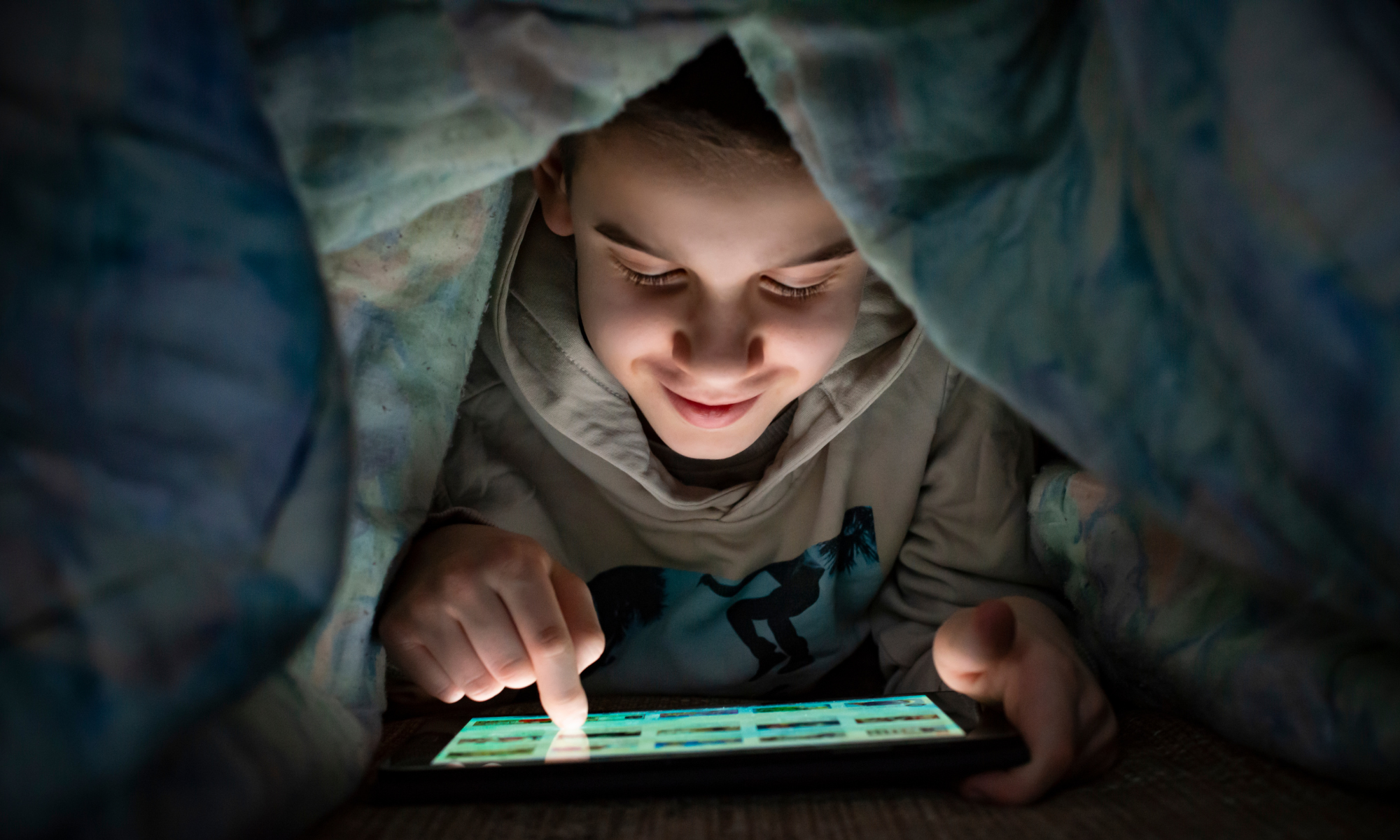 Fotografia di un bambino che legge su tablet sotto a una coperta
